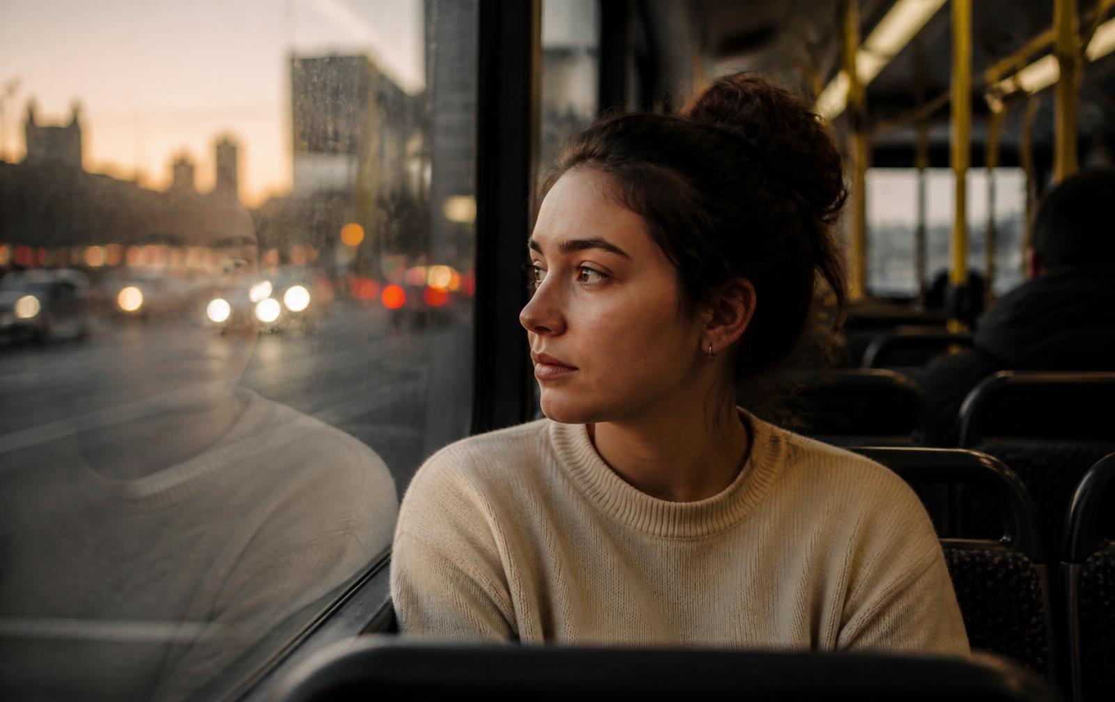 A woman on a bus at dusk, her reflection in the window beside her